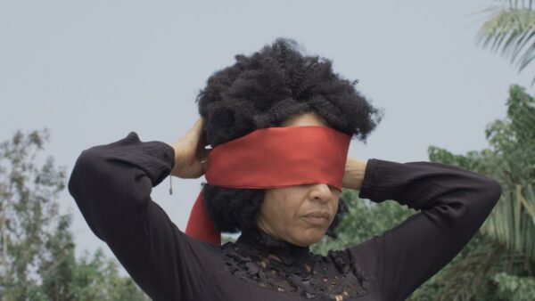 A still image from a film by Mónica de Miranda featuring a figure tying a red blindfold over their eyes.